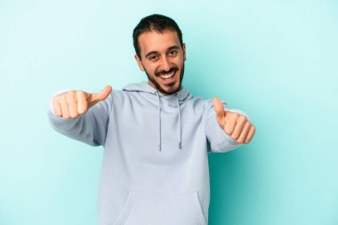 Kafkasyalı genç adam mavi arka planda izole edilmiş. Baş parmağını kaldırıyor, gülümsüyor ve kendinden emin..