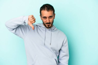 Mavi arka planda izole edilmiş genç beyaz bir adam baş parmağını aşağı gösteriyor ve hoşlanmadığını ifade ediyor..