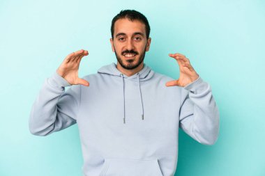 Kafkasyalı genç adam mavi arka planda izole edilmiş. Elinde avuç içi olan bir şey var ve kameraya sunuyor..