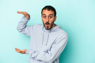 Mavi arka planda izole genç beyaz adam şok ve hayret içinde elleri arasında bir kopya boşluk tutuyor.