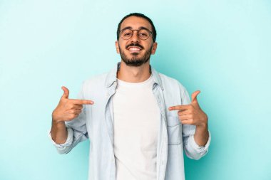 Mavi arka planda izole edilmiş genç beyaz adam bir gömlek fotokopi alanını işaret ediyor, gururlu ve kendinden emin.