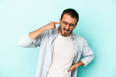 Mavi arka planda izole edilmiş genç beyaz adam kulaklarını elleriyle kapatıyor..