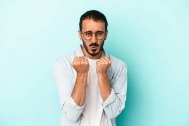 Mavi arka planda izole edilmiş genç beyaz adam yumruğunu kameraya gösteriyor, agresif yüz ifadesi var..