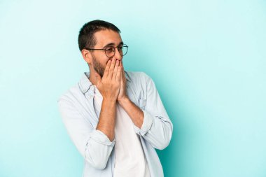 Mavi arka planda izole edilmiş genç beyaz bir adam bir şeye gülüyor, ağızlarını ellerle kapatıyor..