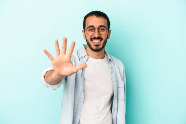 Mavi arka planda izole edilmiş genç beyaz adam gülümseyerek beş numarayı parmaklarıyla gösteriyor..