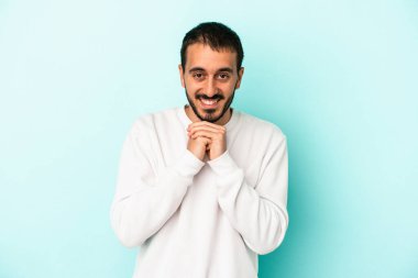 Mavi arka planda izole edilmiş genç beyaz adam ellerini çenesinin altında tutuyor, mutlu bir şekilde yana bakıyor..