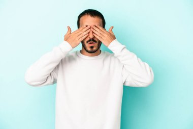 Mavi arka planda izole edilmiş genç beyaz adam elleriyle gözlerini kapatmaktan korkuyor..