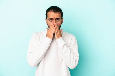 Mavi arka planda izole edilmiş genç beyaz adam şok olmuş, ağzı ellerle kaplanmış, yeni bir şey keşfetmeye hevesli..
