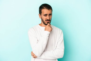 Kafkasyalı genç adam mavi arka planda izole edilmiş düşünüyor ve bakıyor, yansıtıyor, düşünüyor, bir fantezi kuruyor..