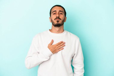 Kafkasyalı genç adam mavi arka planda izole edilmiş yemin ediyor, elini göğsüne koyuyor..