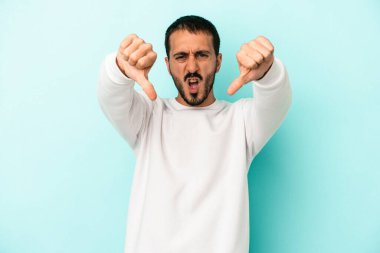 Mavi arka planda izole edilmiş genç beyaz bir adam baş parmağını aşağı gösteriyor ve hoşlanmadığını ifade ediyor..