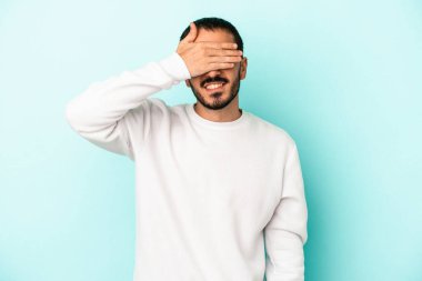 Mavi arka planda soyutlanmış genç beyaz bir adam elleriyle gözlerini kapatıyor, geniş bir gülümsemeyle bir sürpriz bekliyor..