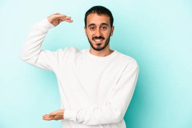 Kafkasyalı genç adam mavi arka planda izole edilmiş. Elinde işaret parmaklarıyla küçük bir şey tutuyor, gülümsüyor ve kendinden emin..