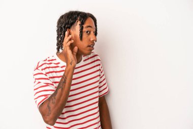Genç Afrikalı Amerikalı adam beyaz arka planda izole edilmiş bir şekilde bir dedikoduyu dinlemeye çalışıyor..