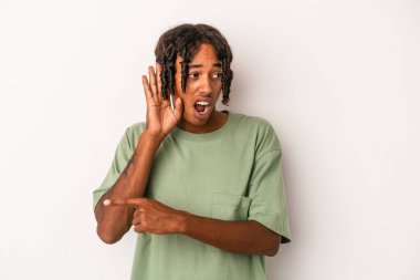 Genç Afrikalı Amerikalı adam beyaz arka planda izole edilmiş bir şekilde bir dedikoduyu dinlemeye çalışıyor..