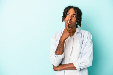 Genç Afrikalı Amerikalı bir adam mavi arka planda izole edilmiş şüpheli ve şüpheci bir ifadeyle yana bakıyor..
