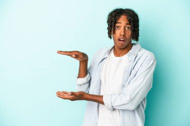 Genç Afrikalı Amerikalı bir adam mavi arka planda izole edilmiş. Elleri arasında bir kopya alanı tutarken şok olmuş ve hayrete düşmüştü..