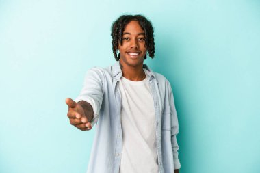 Genç Afro-Amerikan adam mavi arka planda izole edildi. Kameraya doğru elini uzatarak selamlama hareketi yapıyor..