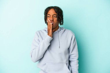 Genç Afrikalı Amerikalı bir adam mavi arka planda izole edilmiş esniyor ve ağzı eliyle kapalı yorgun bir jest yapıyor..