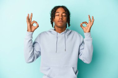 Genç Afrikalı Amerikalı erkek mavi arka planda izole edilmiş yoğun bir günün ardından rahatlıyor, yoga yapıyor..