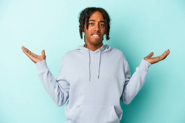 Genç Afrikalı Amerikalı bir adam mavi arka planda izole edilmiş. Kafası karışmış ve omuz silkiyor..