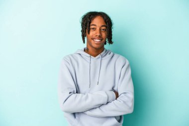 Genç Afro-Amerikalı, mavi arka planda izole edilmiş, kendinden emin, kararlılıkla kollarını kavuşturuyor..