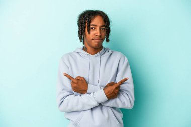 Genç Afrikalı Amerikalı adam mavi arka planda iki seçenek arasında seçim yapmaya çalışıyor..