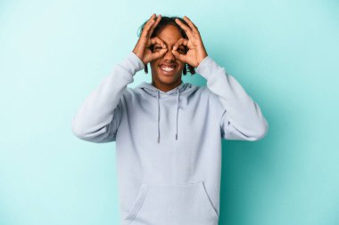 Genç Afro-Amerikalı adam mavi arka planda izole edilmiş, gözlerinde sorun yok işareti var.