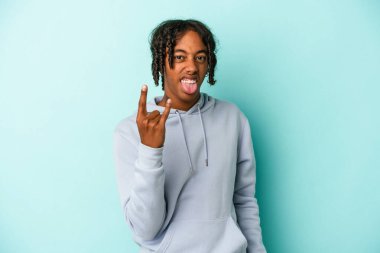 Genç Afrikalı Amerikalı adam mavi arka planda izole edilmiş. Parmaklarıyla kaya hareketi gösteriyor.