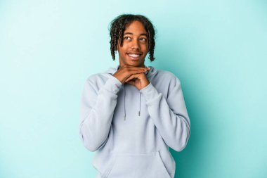 Mavi arka planda izole edilmiş genç Afrikalı Amerikalı ellerini çenesinin altında tutuyor ve mutlu bir şekilde yan tarafa bakıyor..