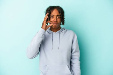 Genç Afrikalı Amerikalı bir adam mavi arka planda izole edilmiş ve parmakları dudaklarında bir sır saklıyor..