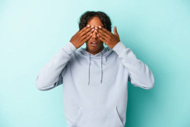 Genç Afrikalı Amerikalı bir adam mavi arka planda izole edilmiş. Gözleri elleriyle kapatmaktan korkuyor..