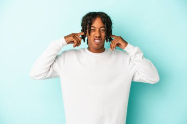 Genç Afrikalı Amerikalı bir adam mavi arka planda izole edilmiş parmaklarıyla kulaklarını kapatıyor, gürültülü bir ortam yüzünden stresli ve çaresiz..
