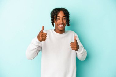 Genç Afrikalı Amerikalı adam mavi arka planda izole edilmiş. Baş parmağını kaldırıyor, gülümsüyor ve kendinden emin..