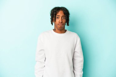 Genç Afrikalı Amerikalı bir adam mavi arka planda izole edilmiş omuzlarını silker ve gözleri açık kalır..