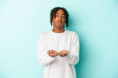 Genç Afrikalı Amerikalı adam mavi arka planda izole edilmiş. Elinde avuç içi olan bir şey var. Kameraya teklif ediyor..