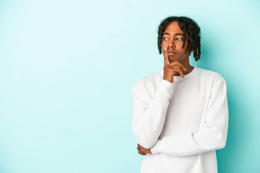 Genç Afrikalı Amerikalı bir adam mavi arka planda izole edilmiş şüpheli ve şüpheci bir ifadeyle yana bakıyor..