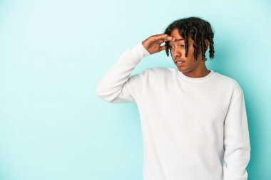 Genç Afrikalı Amerikalı adam mavi arka planda izole edilmiş, uzaklara bakıyor ve elini alnına koyuyor..