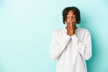 Genç Afrikalı Amerikalı bir adam mavi arka planda izole edilmiş. Düşünceli bir şekilde bir fotokopi odasına bakıyor. Ağzını eliyle kapatıyor..