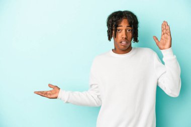 Mavi arka planda izole edilmiş genç Afro-Amerikalı bir adam avuç içinde fotokopi alanı tutuyor, el üstünde tutuyor. Şaşırmış ve mutlu..