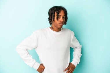 Mavi arka planda izole edilmiş, hoşnutsuzca kaşlarını çatmış, kollarını kavuşturmuş genç Afrikalı Amerikalı bir adam..