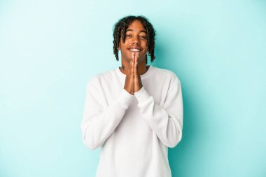 Mavi arka planda izole edilmiş genç Afro-Amerikan bir adam el ele tutuşup dua ederken kendine güveniyor..