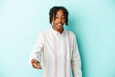 Genç Afro-Amerikan adam mavi arka planda izole edildi. Kameraya doğru elini uzatarak selamlama hareketi yapıyor..