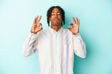 Genç Afrikalı Amerikalı erkek mavi arka planda izole edilmiş yoğun bir günün ardından rahatlıyor, yoga yapıyor..