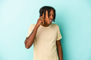 Genç Afrikalı Amerikalı bir adam mavi arka planda izole edilmiş parmaklarıyla kulaklarını kapatıyor, gürültülü bir ortam yüzünden stresli ve çaresiz..