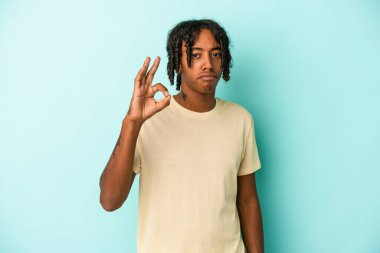 Genç Afrikalı Amerikalı bir adam mavi arka planda izole edilmiş bir şekilde bir göz kırpıyor ve elleriyle iyi bir jest yapıyor..