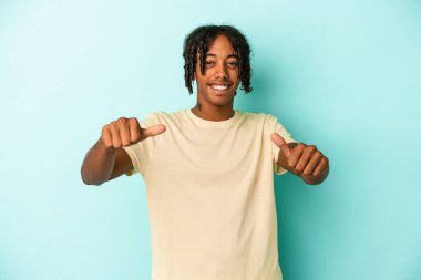 Genç Afrikalı Amerikalı adam mavi arka planda izole edilmiş. Baş parmağını kaldırıyor, gülümsüyor ve kendinden emin..