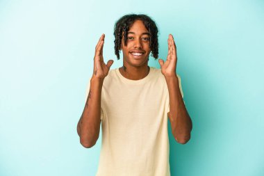 Mavi arka planda izole edilmiş genç Afro-Amerikalı adam yüksek sesle gülüyor ve elini göğsüne koyuyor..