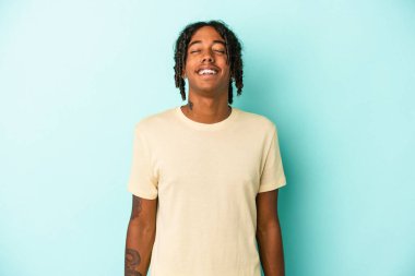 Genç Afrikalı Amerikalı adam mavi arka planda izole edilmiş rahat ve mutlu bir şekilde gülüyor, boynunda dişlerini gösteriyor..