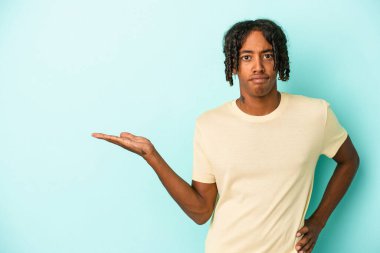 Mavi arka planda izole edilmiş genç Afrikalı Amerikalı bir adam bir avuç içinde fotokopi alanı ve belinde bir el daha gösteriyor..